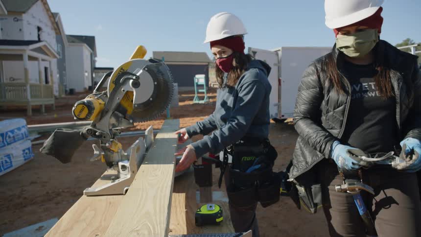 Two women work with saw and wooden board outdoors at construction site