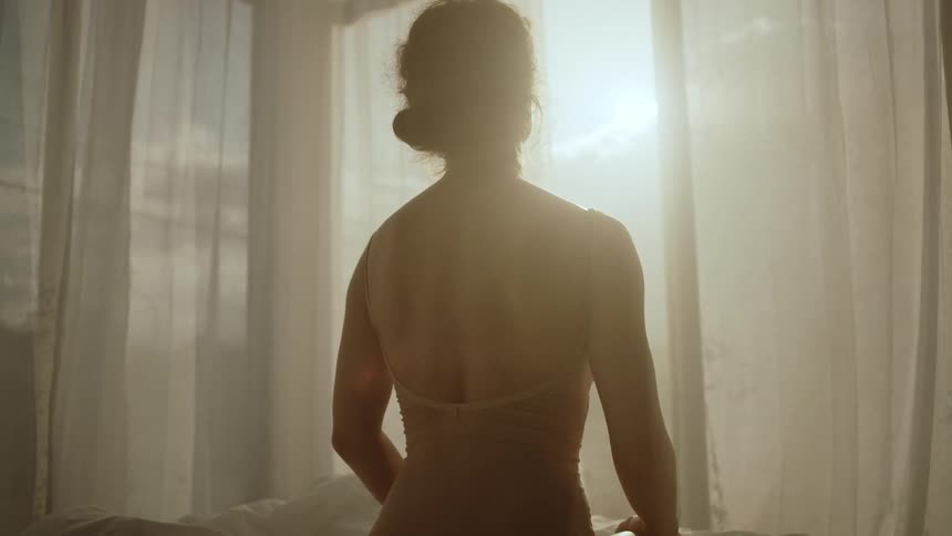 A woman in a corset stands and looks out her bedroom window