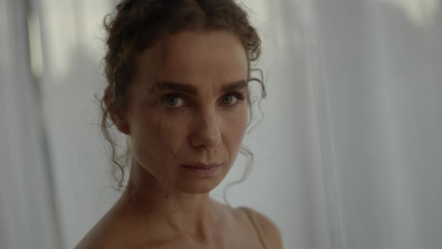 A woman is standing in a room with a white curtain behind her and she has a sad look on her face as she looks up