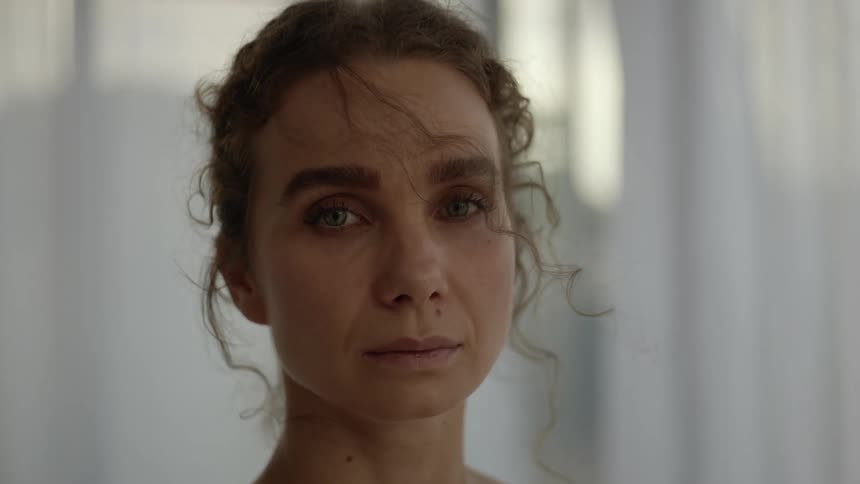 A woman is standing with a white curtain behind her and she has a sad look on her face as her hair moves in the breeze