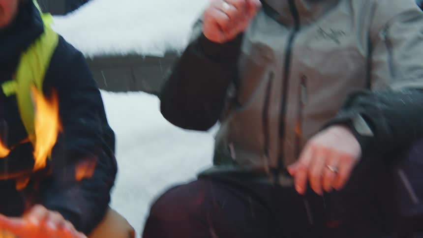 Two friends sitting outside while it snows warm their hands over a fire and then fist bump one another