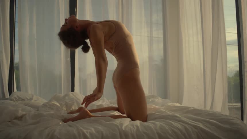 A woman stretches and dances on a bed with a curtain sheer blowing in on her