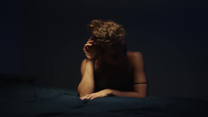 A woman lying on a bed looks around and runs her hand over her face and then lies her head down and runs her hand over the sheets