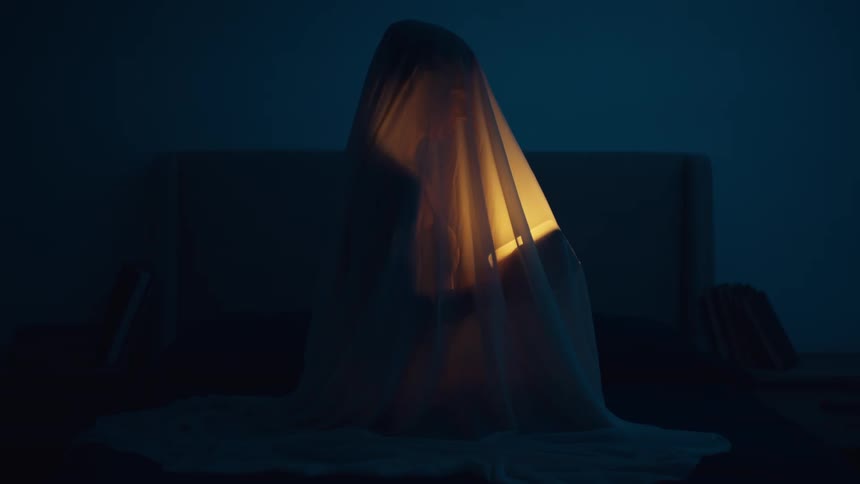 Woman sitting on the bed covered with a bedsheet turns on a flashlight to read a book