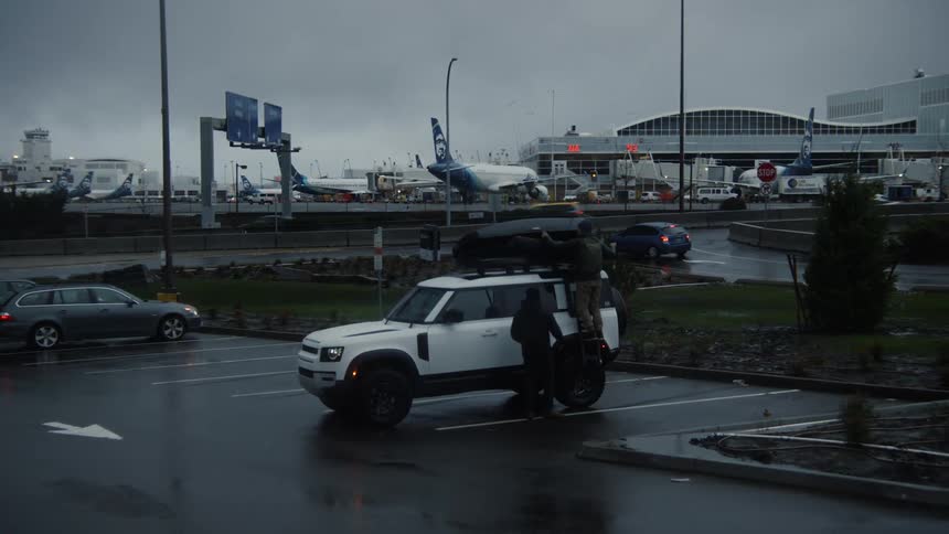 A person standing by a vehicle in a parking lot hands a person standing on the back of the vehicle a large item that they place in a storage case on top of the car