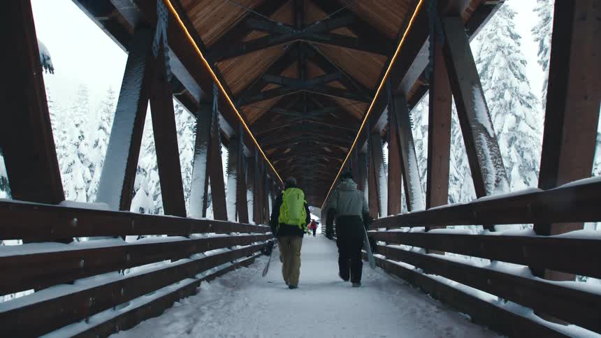 A pair of snowboarders carry their boards across a snowy wooden bridge