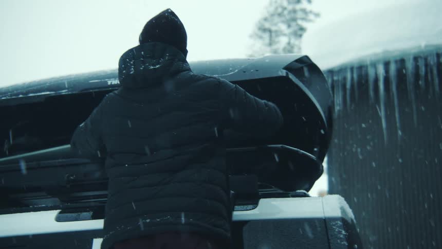 One person hands another person a snowboard to pack into their vehicles rooftop cargo carrier