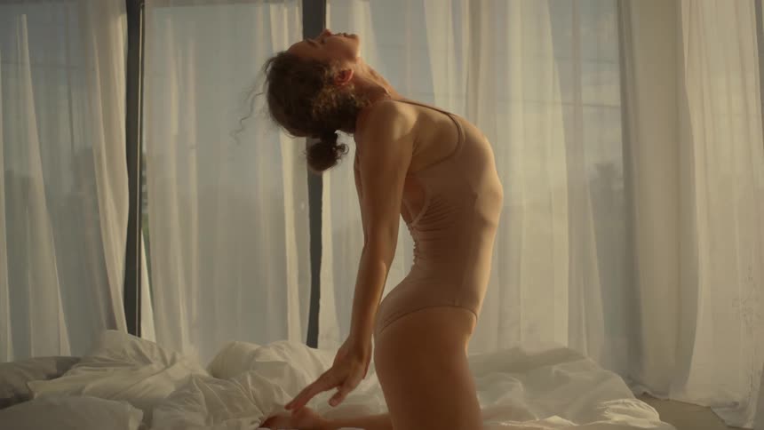 A woman is kneeling on a bed with a canopy around it and moving her arms and arching her back in a dancing motion