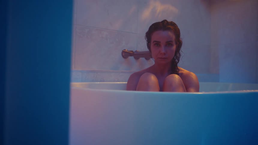 A woman is sitting in a bathtub with her arms around her legs and she looks up with a sad look on her face