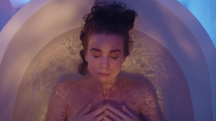 A woman is lying on her back in a bathtub with her hands on her chest and she slides down keeping her face just above the water