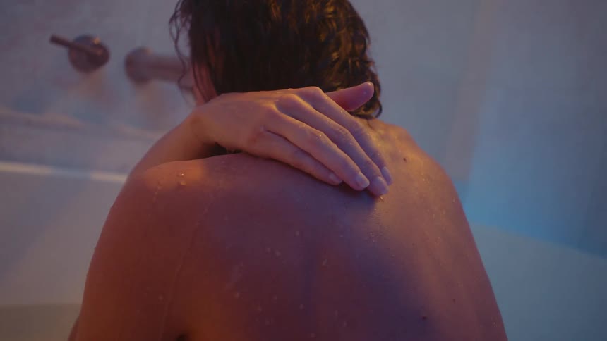 A woman sitting in a bathtub gently pours a handful of water over her shoulder onto her own back