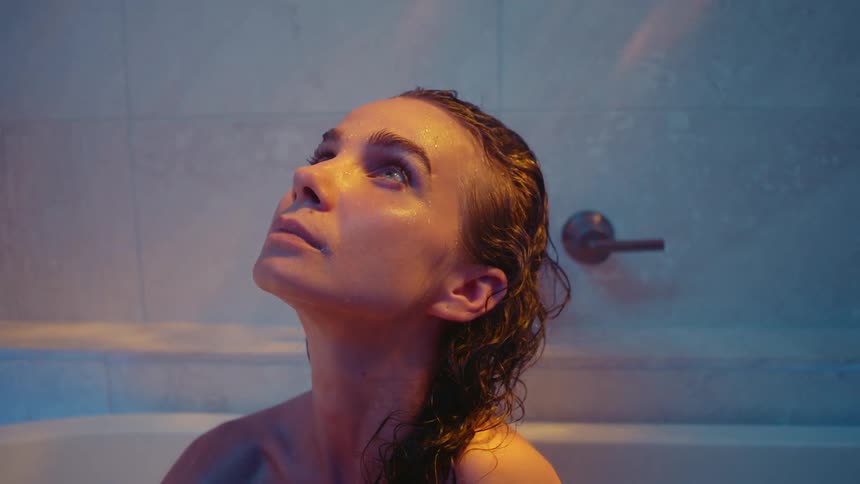 There is a young white woman sitting in a bath tub and looking at the ceiling