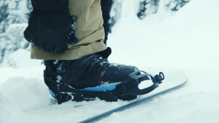 Man carrying a backpack puts on a snowboard atop a mountain covered in white snow