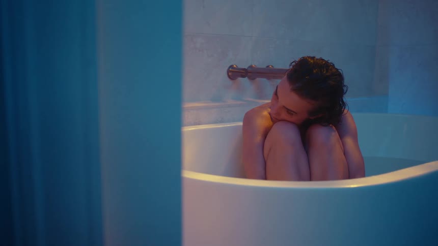 A woman is sitting in a bathtub with her arms around her legs and resting her head on her knees