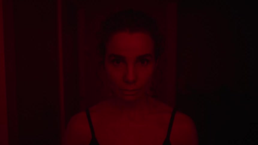 A woman is standing in a room illuminated with red light looking down and then she looks up