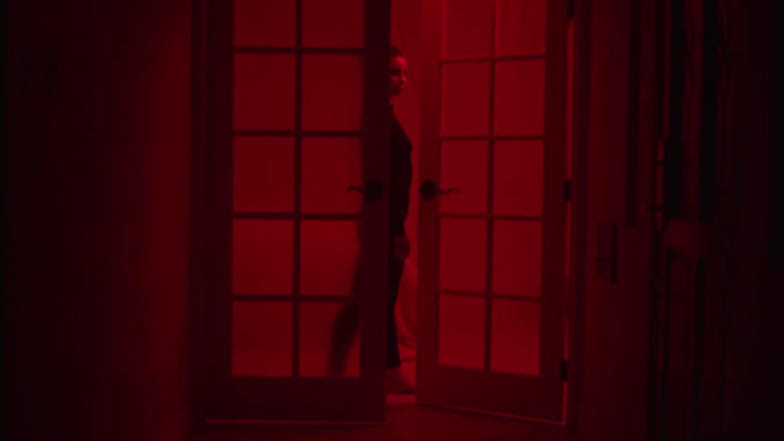 Woman walks forward and shuts door in room lit by red lighting