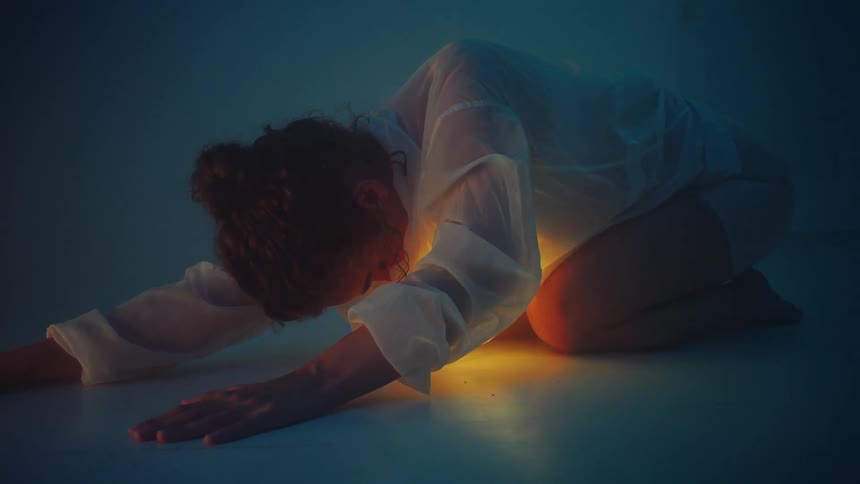A woman in a white shirt is curled up on the floor with a pulsing light beneath her