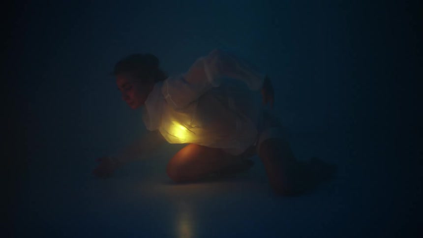 A woman is on her knees moving her arms in a dancing motion in the darkness with a glowing light inside her shirt