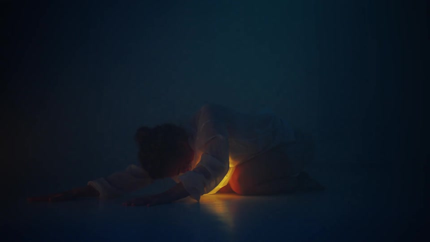 A dancer kneeling on the floor with head bowed and light glowing from beneath her