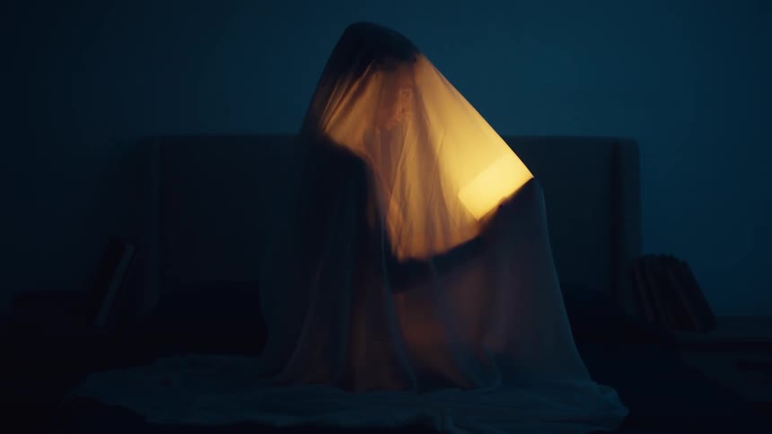 A woman is sitting on a bed covered with a sheet in a dark room and holding a glowing light that illuminates her under the sheet