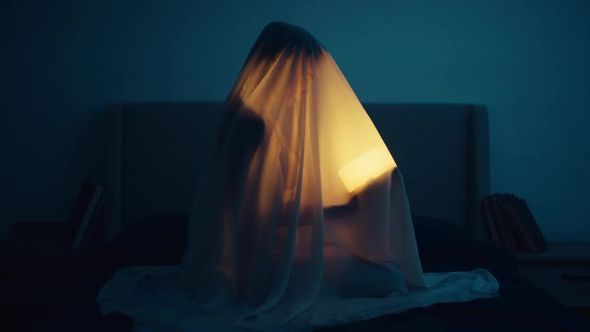 A woman is sitting on a bed covered with a sheet and holding a glowing light illuminates her