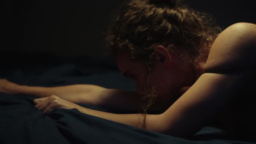 A woman grasps the sheets stretching her body onto the bed from the floor