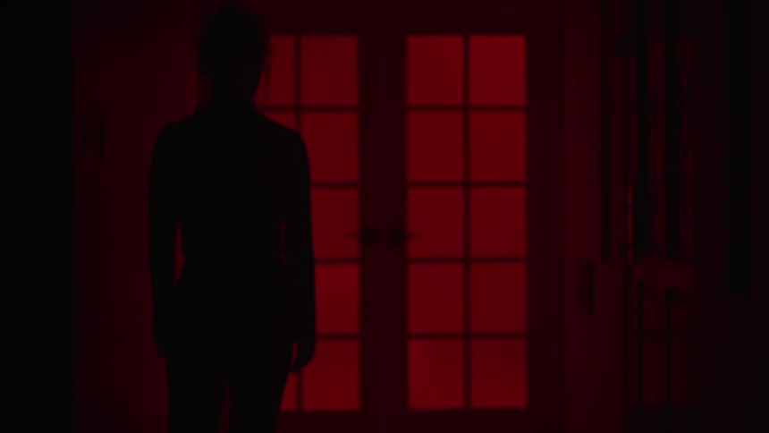 Silhouette of a woman walking to a door in a dark room with red curtains over the windows