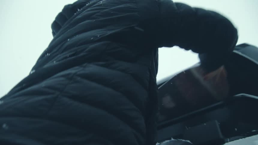 Two people pack snowboard into container on top of vehicle as it snows