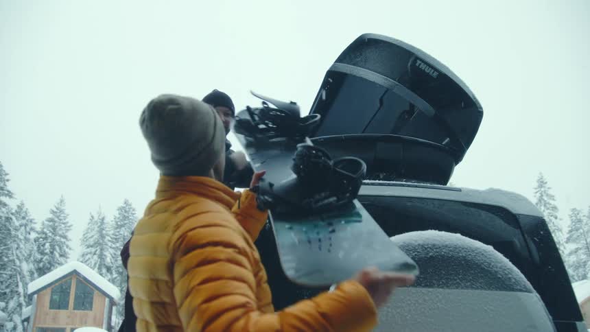 A man hands his friend snowboards and he loads them in to a storage unit on top of a vehicle