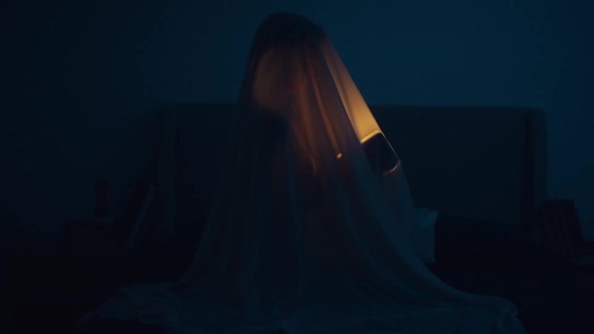 A person with a sheet over themselves turns on a flashlight in the darkness of their room
