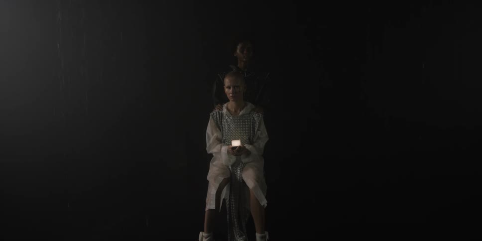 A teenage girl sits on a chair in the dark holding a light as another teenage girl stands behind her and they both turn their heads to look ahead of them
