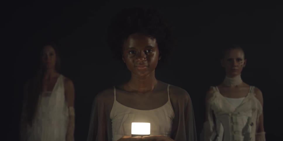 Girl with curly hair and white dress holds lamp smiling at camera in front of two women wearing white dresses in dark room