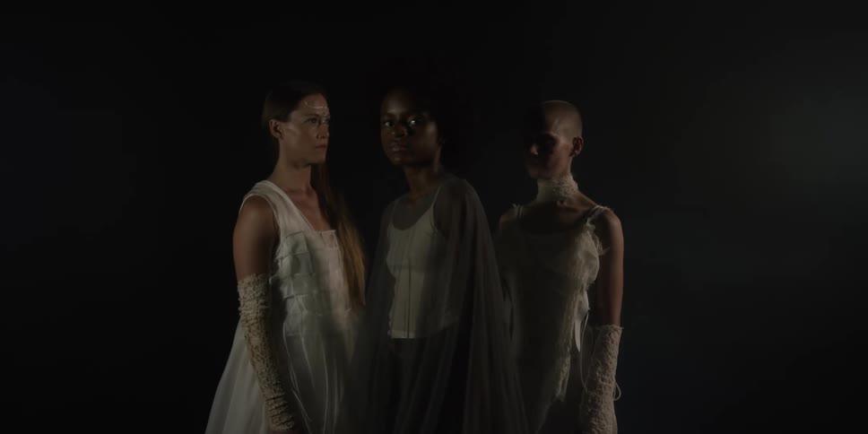 Three women stand facing each other in elaborate gowns before one turns