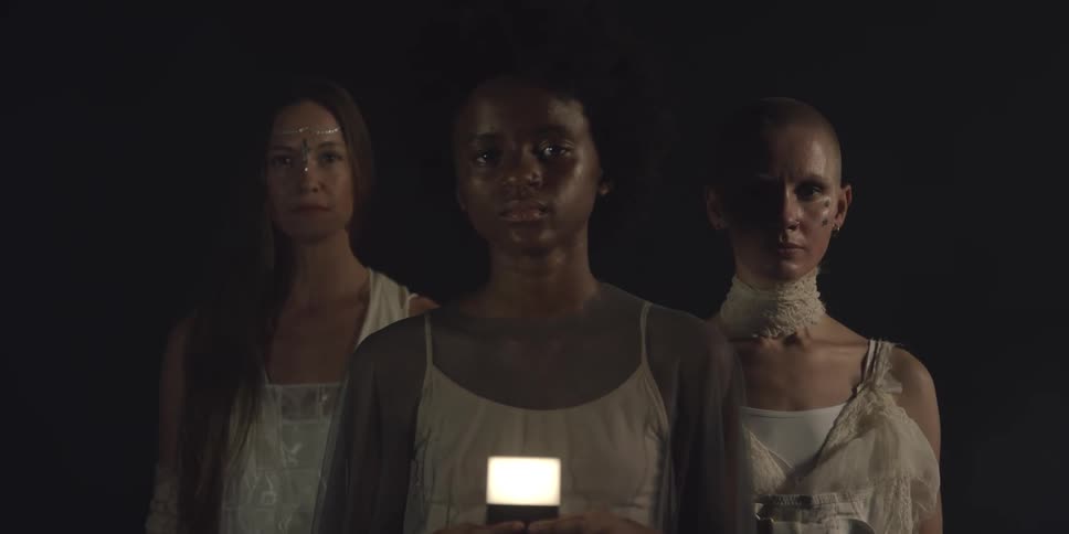 Three women stand in darkness with the one in the front holding a light