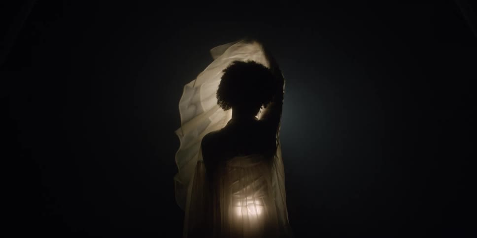 A woman is standing in front of a light in the darkness as she raises her arms the light shines through her cloak