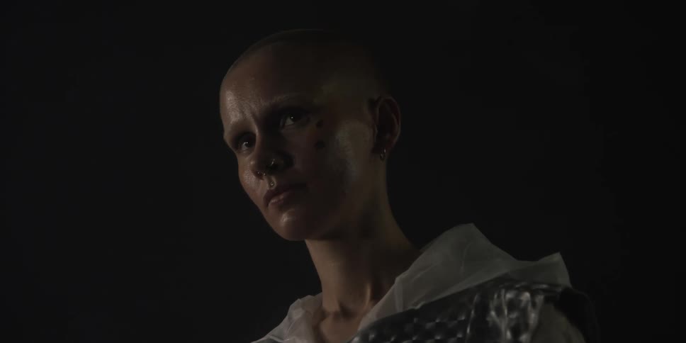 Bald woman with piercings earrings and gray hoodie looks down with sad expression on her face