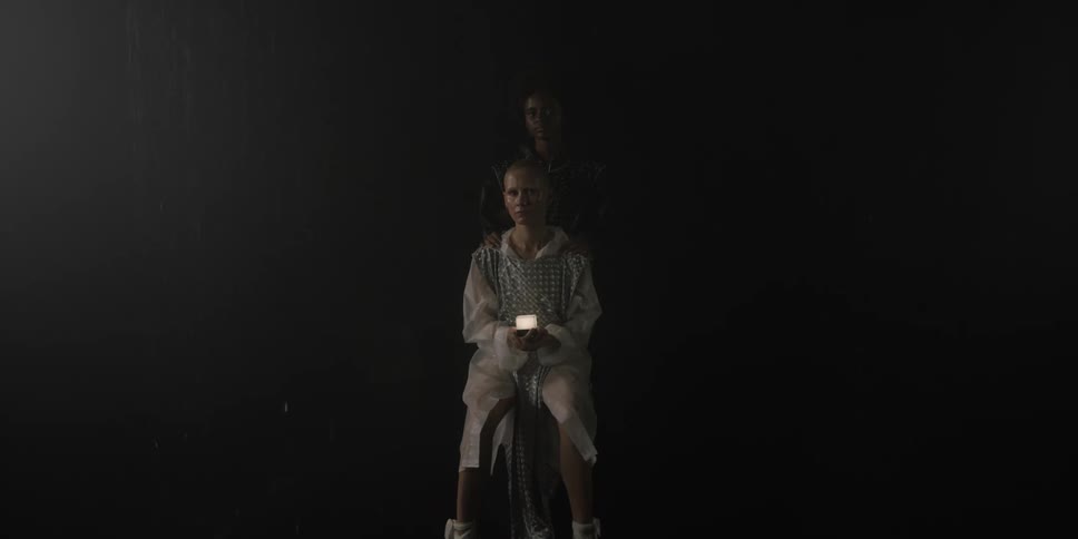 Two female performers one sitting and one standing behind her against black