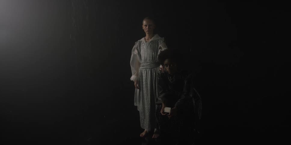 One woman stands while another sits as they pose in dark room while wearing ornate garb