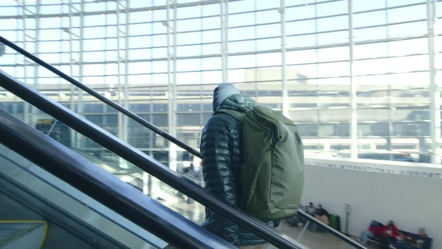 A man wearing a backpack and carrying luggage is riding up and escalator at an airport