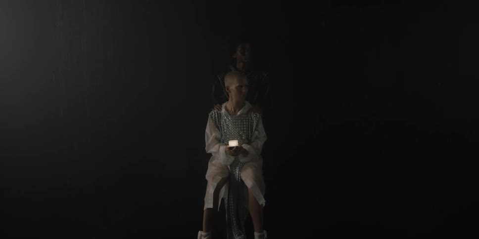 A teenage girl sits in the dark holding a candle and another teen stands behind her and they turn and look to the side