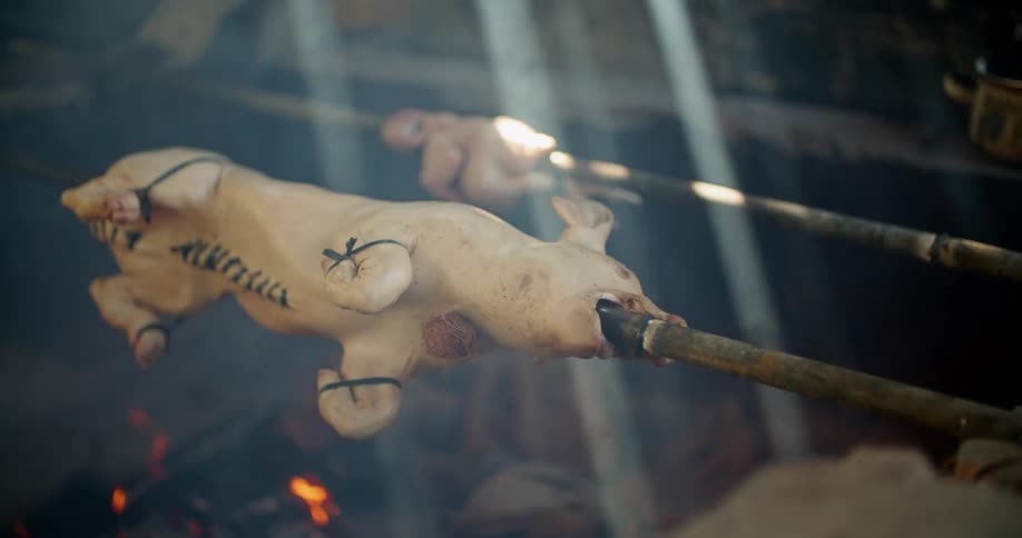 Spit pig and chicken are roasted over flame as light rays pass through roof