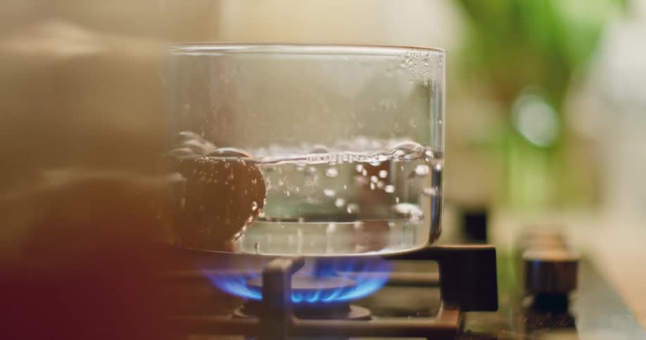 An egg is placed into a glass pot of boiling water on a stove