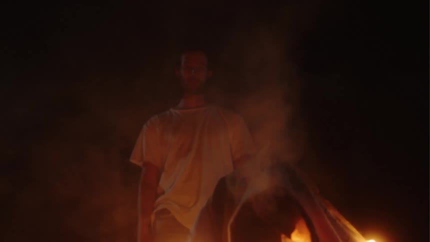 A man stands and stares at the flames of a small fire burning in front of him