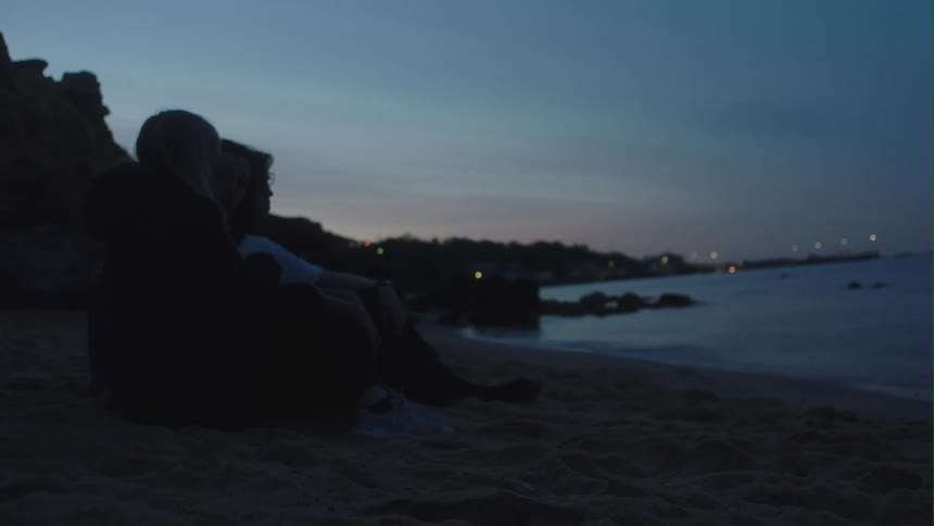 People are sitting together on a beach as the waves roll in and the sun has set