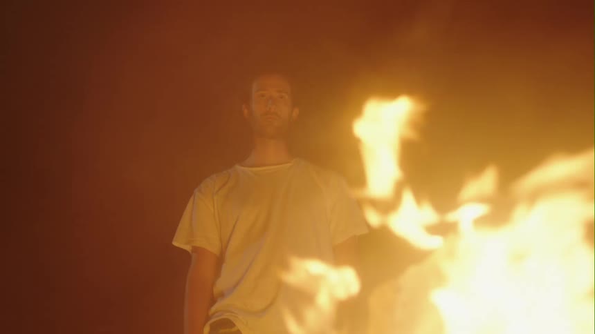 Man with long hair and white t shirt watches bonfire on the beach at night