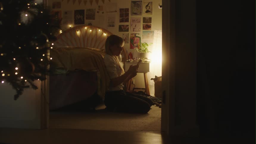 Woman with white shirt and black pants sits next to bed taking selfie with smartphone in dim lit room