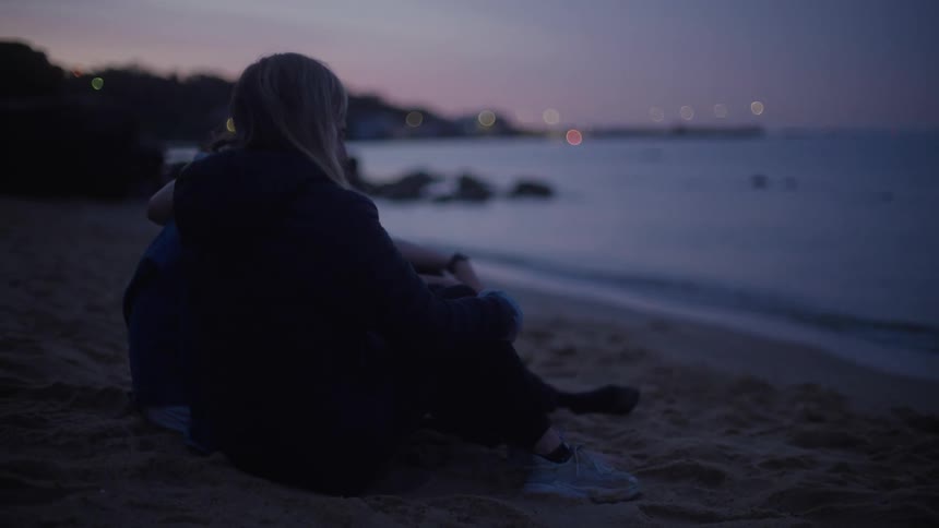 Two men and blonde woman sit on the the beach at sunset next to the sea putting blanket on and cuddling