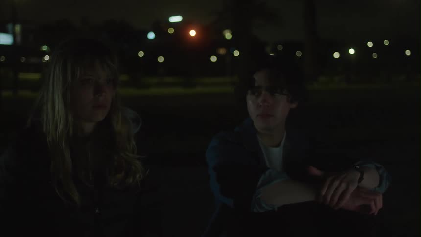 Man sits down on ground next to motionless staring woman and turns to look at her