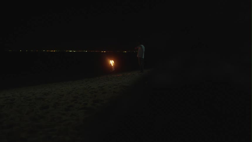 Man with white shirt and shorts stands by bonfire near the sea on the beach at night