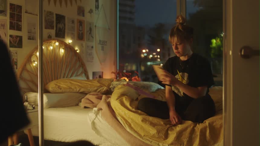 Young woman sitting on the bed in front of a mirror throws away a phone while crying